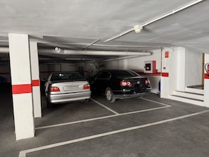 Un parcheggio sotterraneo con due auto parcheggiate in posti adiacenti, pilastri bianchi con strisce rosse e un estintore fissato al muro.
