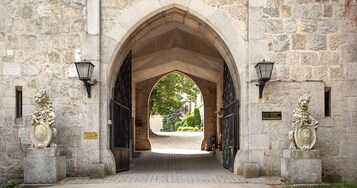 Uma entrada de pedra em arco com duas estátuas de leão de cada lado, um caminho pavimentado e uma vista de vegetação ao fundo.