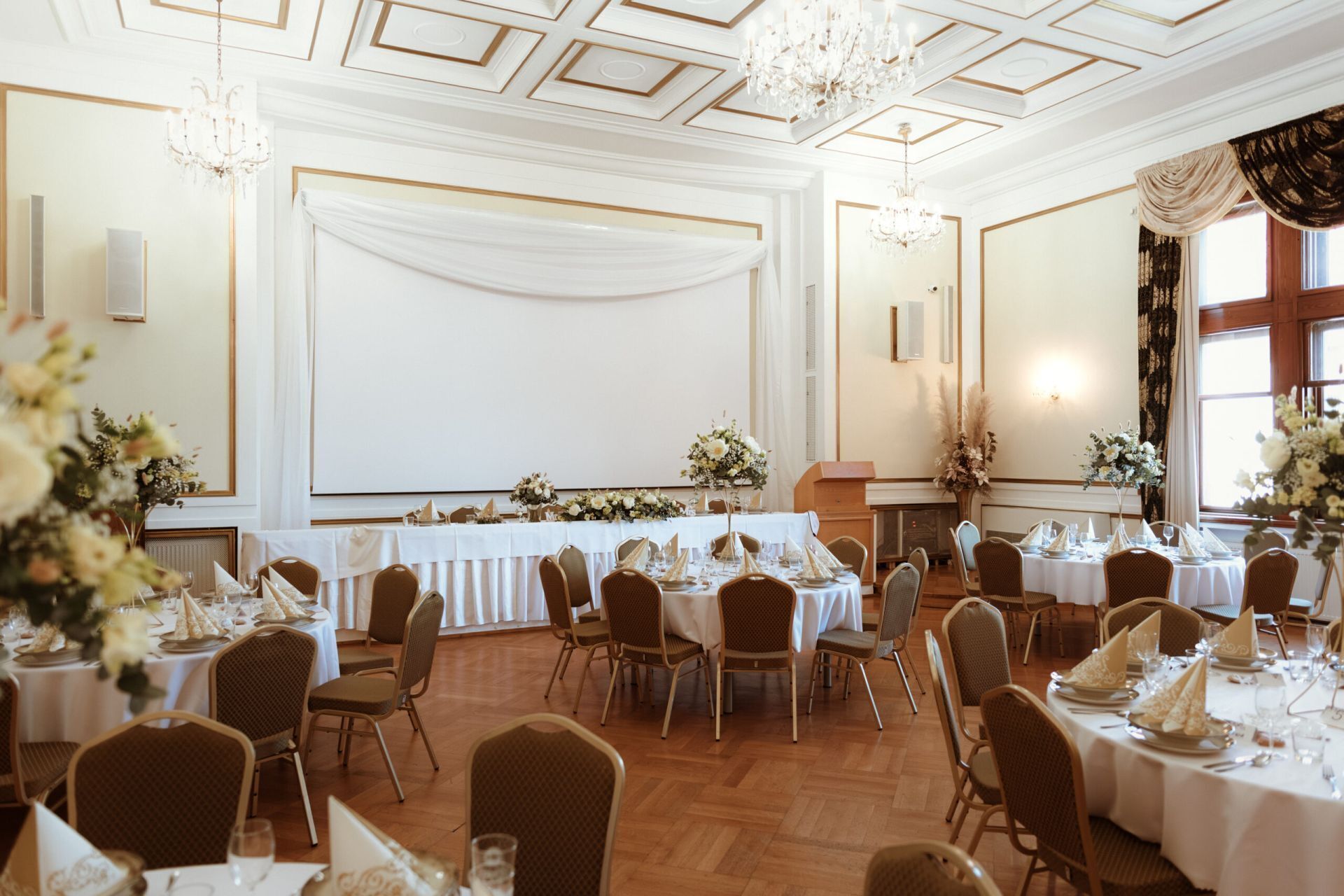 A banquet hall with round tables, chairs, chandeliers, and floral arrangements.