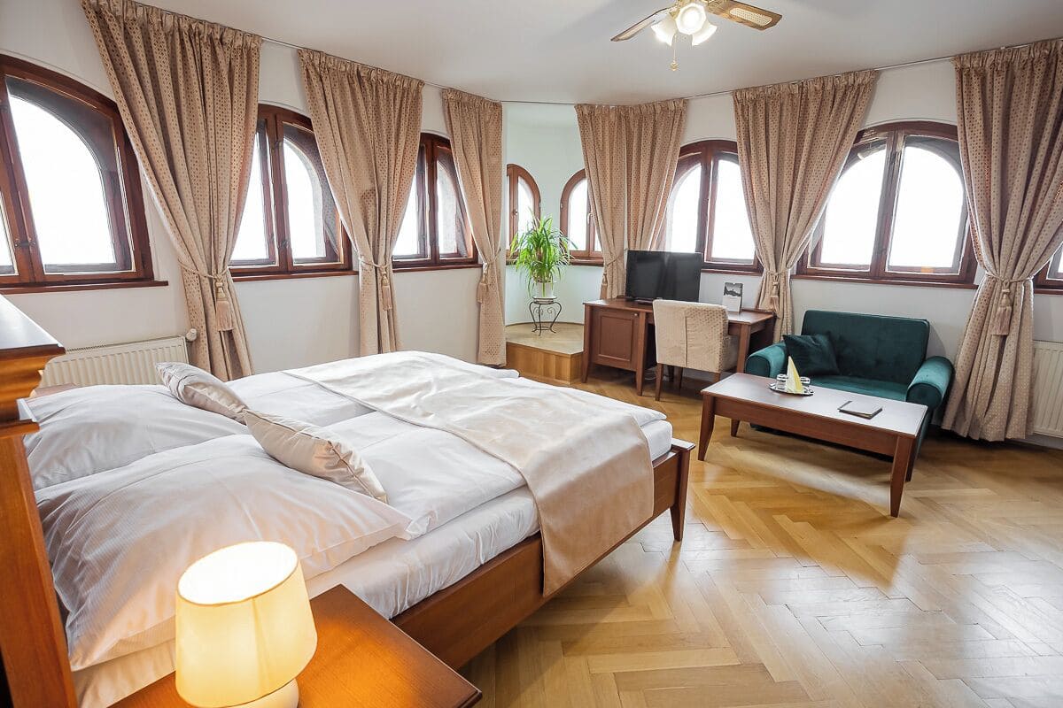 A hotel room with a bed, a desk, a television, a sofa, and large windows with curtains.