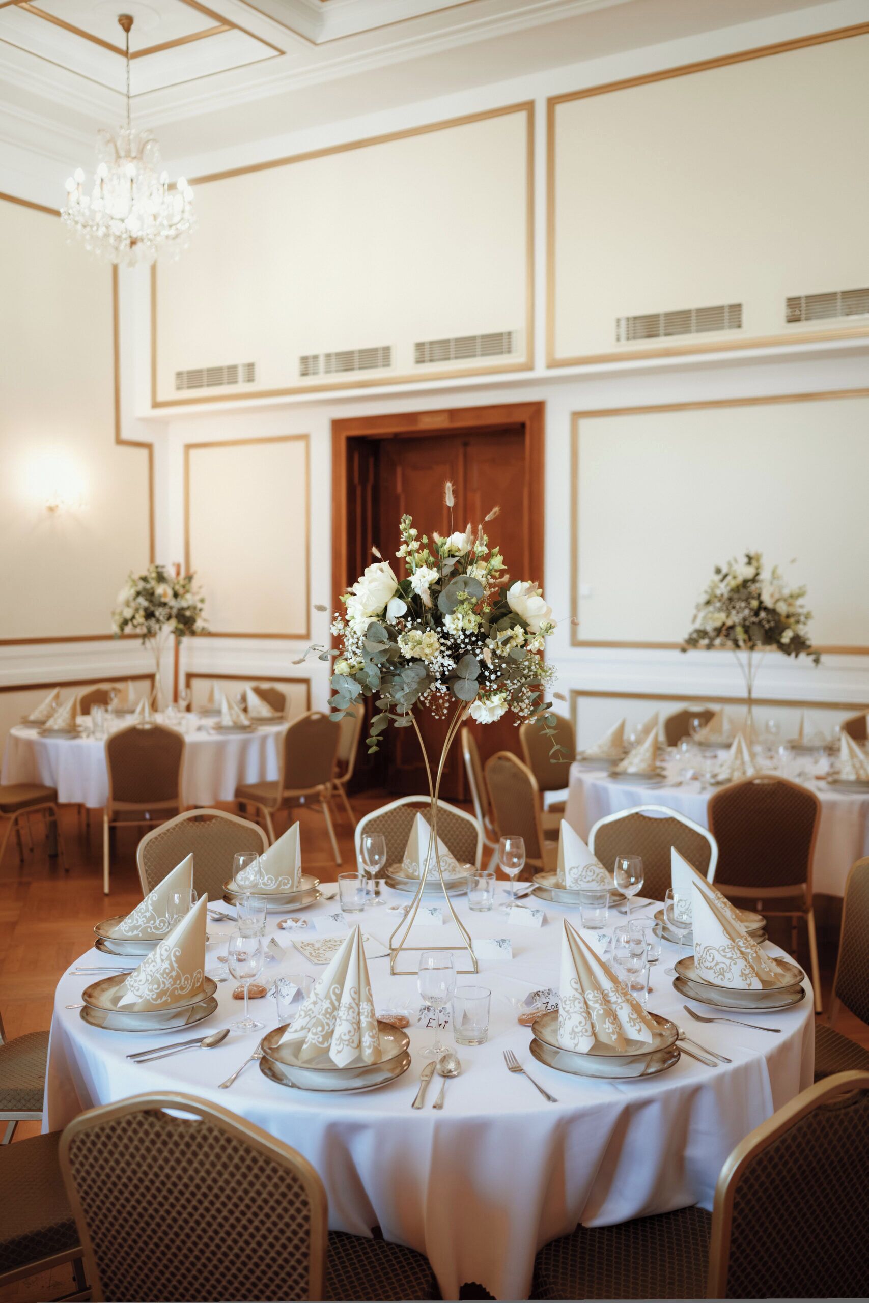 A banquet hall with round tables set for a formal event, featuring white tablecloths, gold accents, and floral centerpieces.
