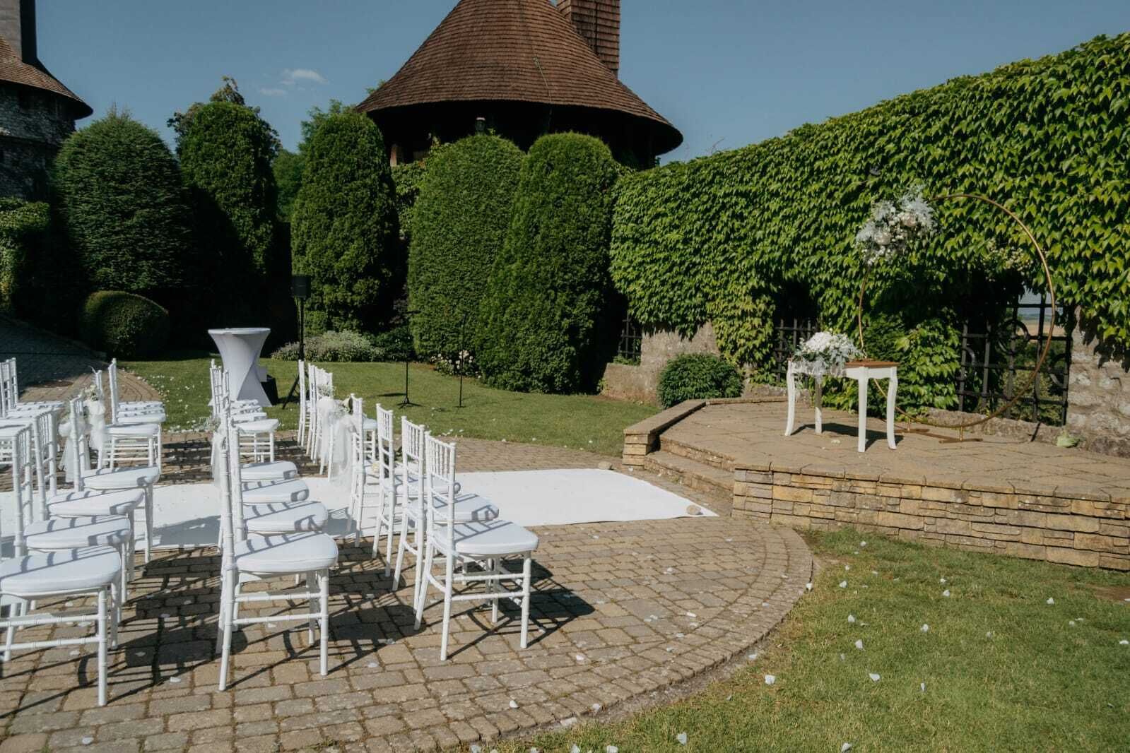 An outdoor wedding setup with white chairs and tables arranged on a paved area, surrounded by greenery and a stone wall.