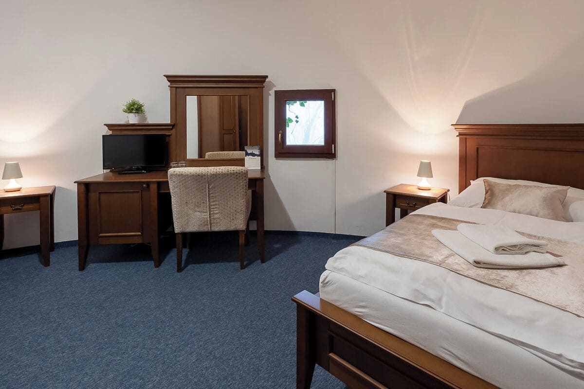 A hotel room with a bed, a desk with a chair, a television, and a small window.
