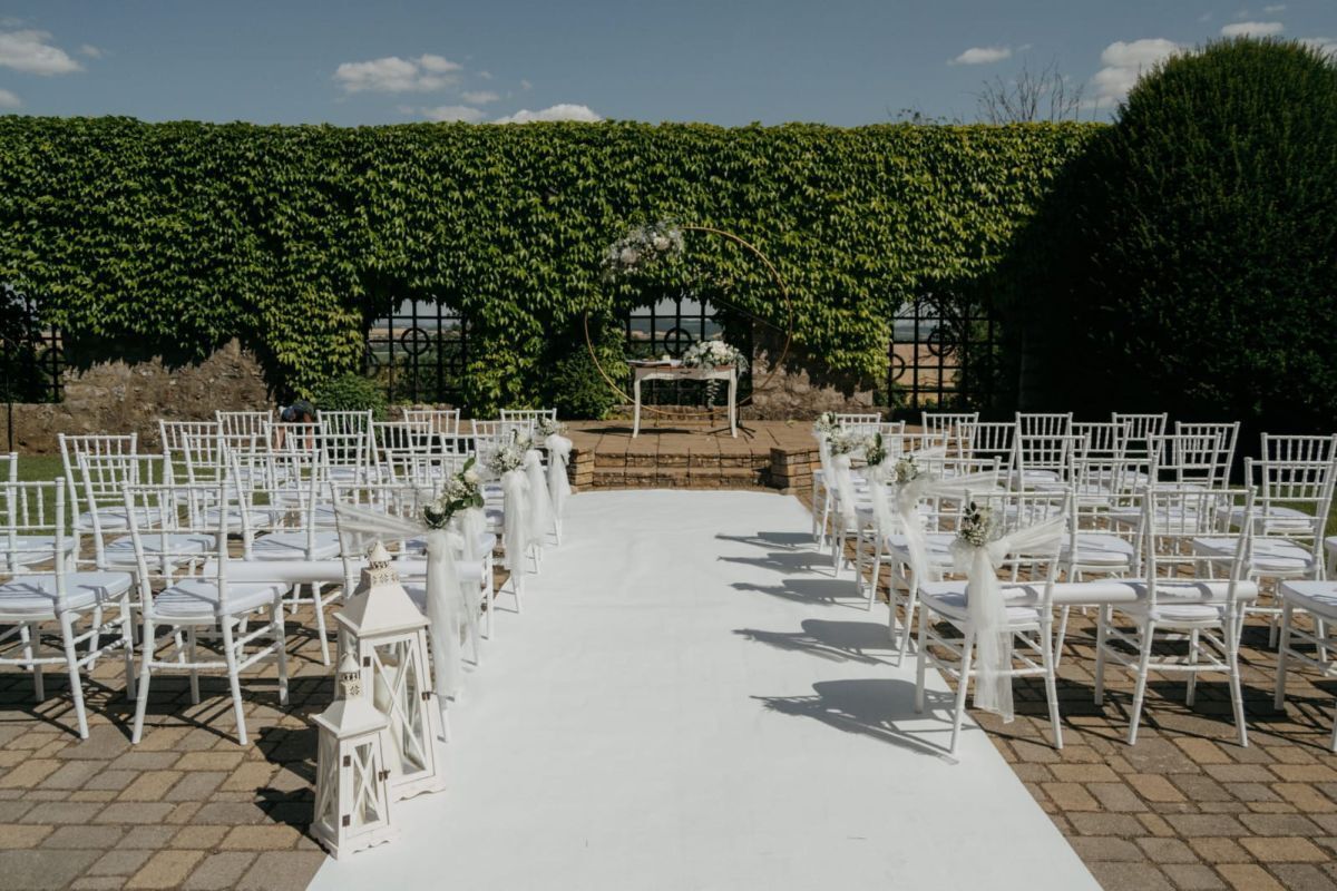 Un lieu de cérémonie de mariage en plein air, avec des chaises blanches disposées en rangées, une allée blanche et un décor de verdure luxuriante.