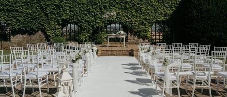 Un espacio para bodas al aire libre con sillas blancas dispuestas en filas, un pasillo blanco y un fondo de follaje verde.