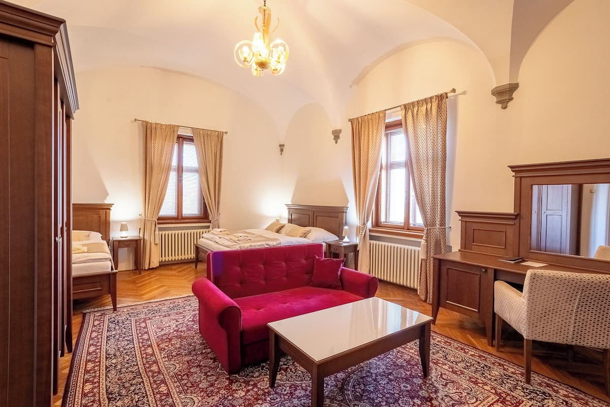 A hotel room with a bed, a red sofa, a small table, a chair, a desk, and a mirror.