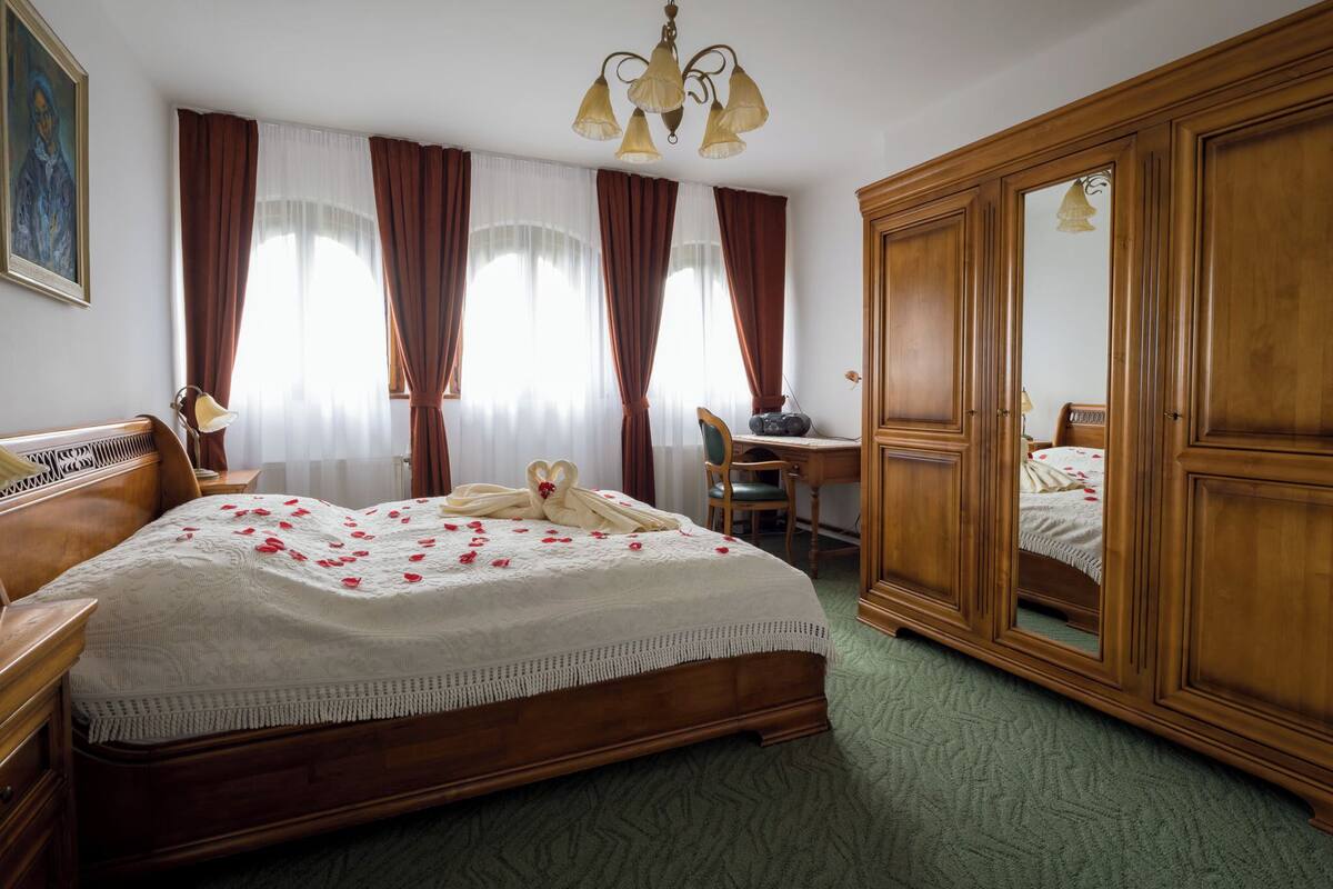 A hotel room with a large bed, wooden wardrobe, a desk with a chair, and a painting on the wall.