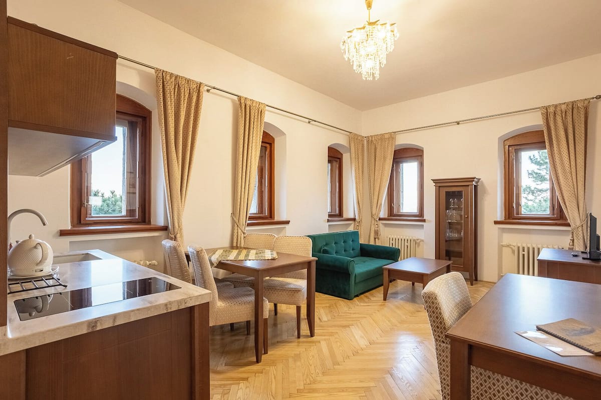 A room with a kitchen area, dining table, chairs, a green sofa, and a chandelier.