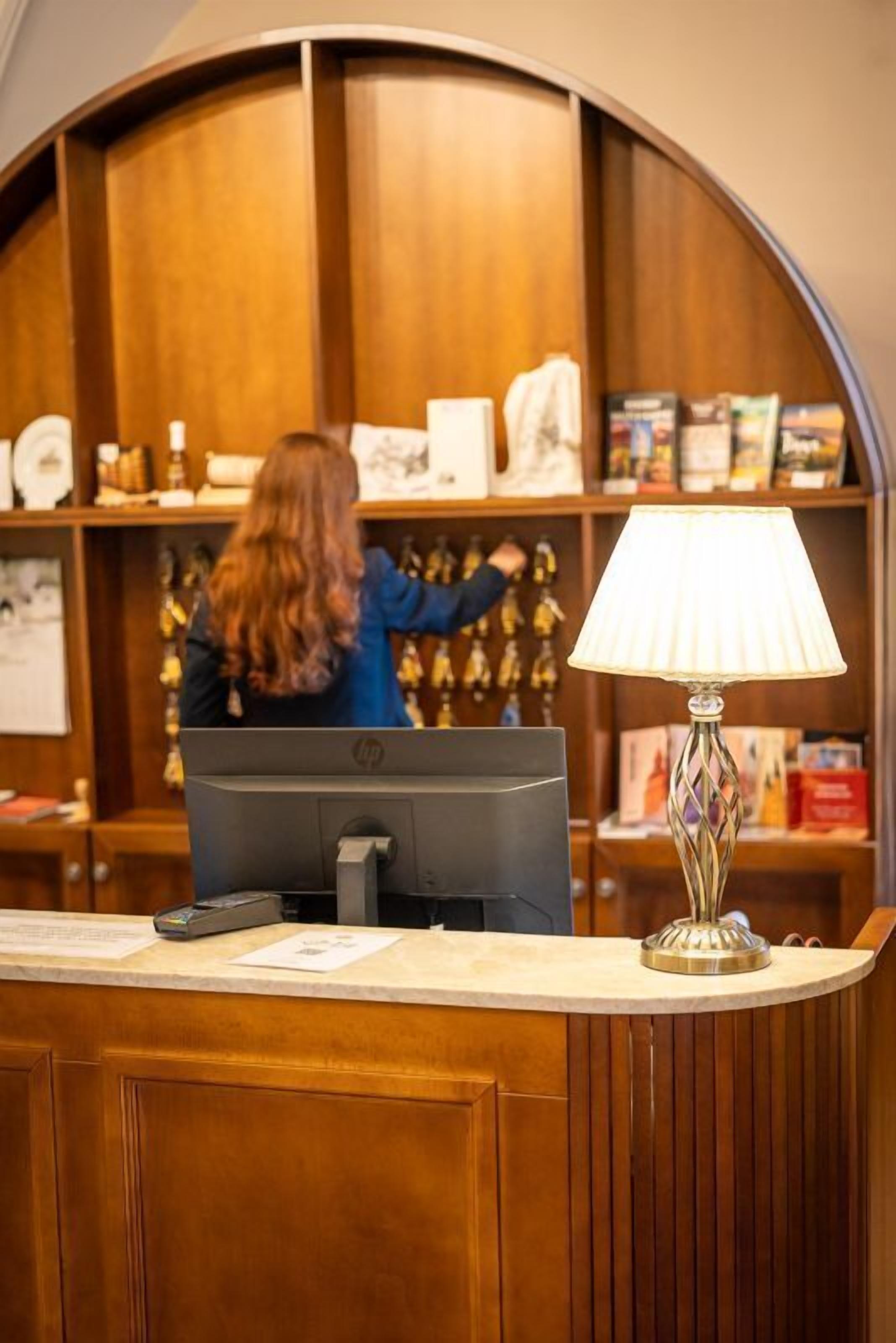 Een receptiebalie met een computer, een lamp en een persoon achter de balie.