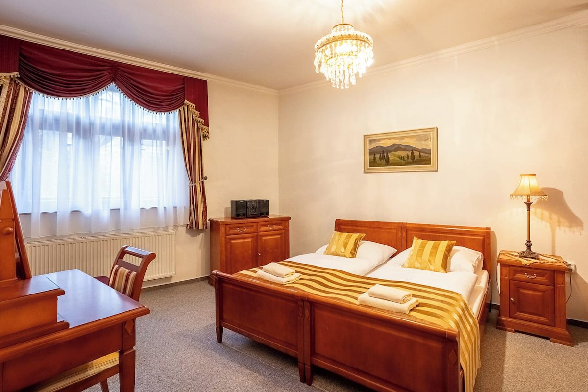 A hotel room with a wooden bed, a desk, a chair, a radiator, a chandelier, a painting, and a bedside table with a lamp.