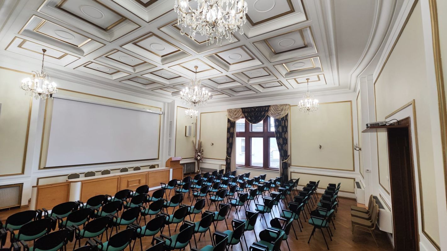 Un salón de conferencias con una pizarra, filas de sillas, un atril y una araña de cristal.