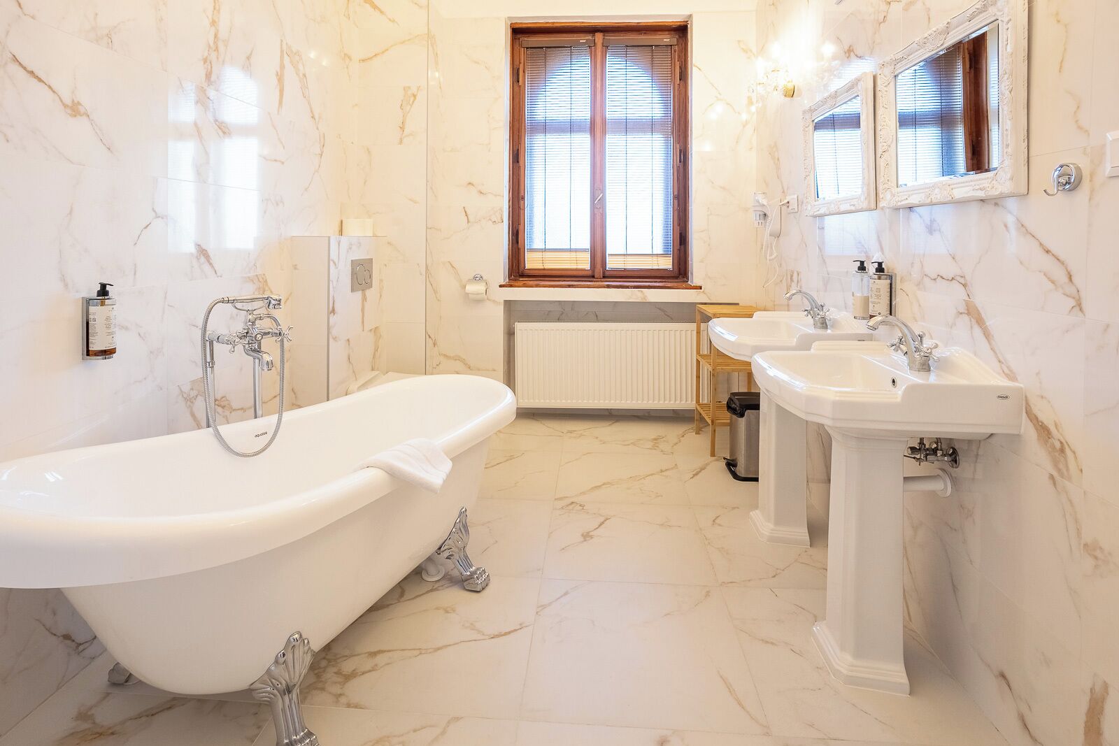 A bathroom with a freestanding bathtub, two sinks, a window with blinds, and marble walls.