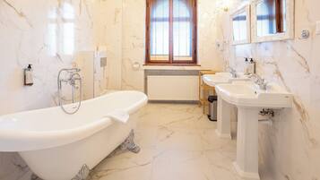 A bathroom with a freestanding bathtub, two sinks, a window with blinds, and marble walls.
