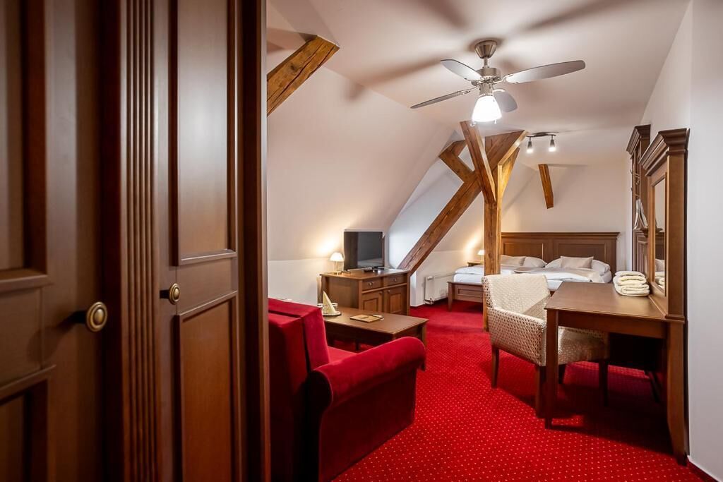 Una habitación acogedora con alfombra roja, una cama, un escritorio, una silla, un televisor y un ventilador de techo.