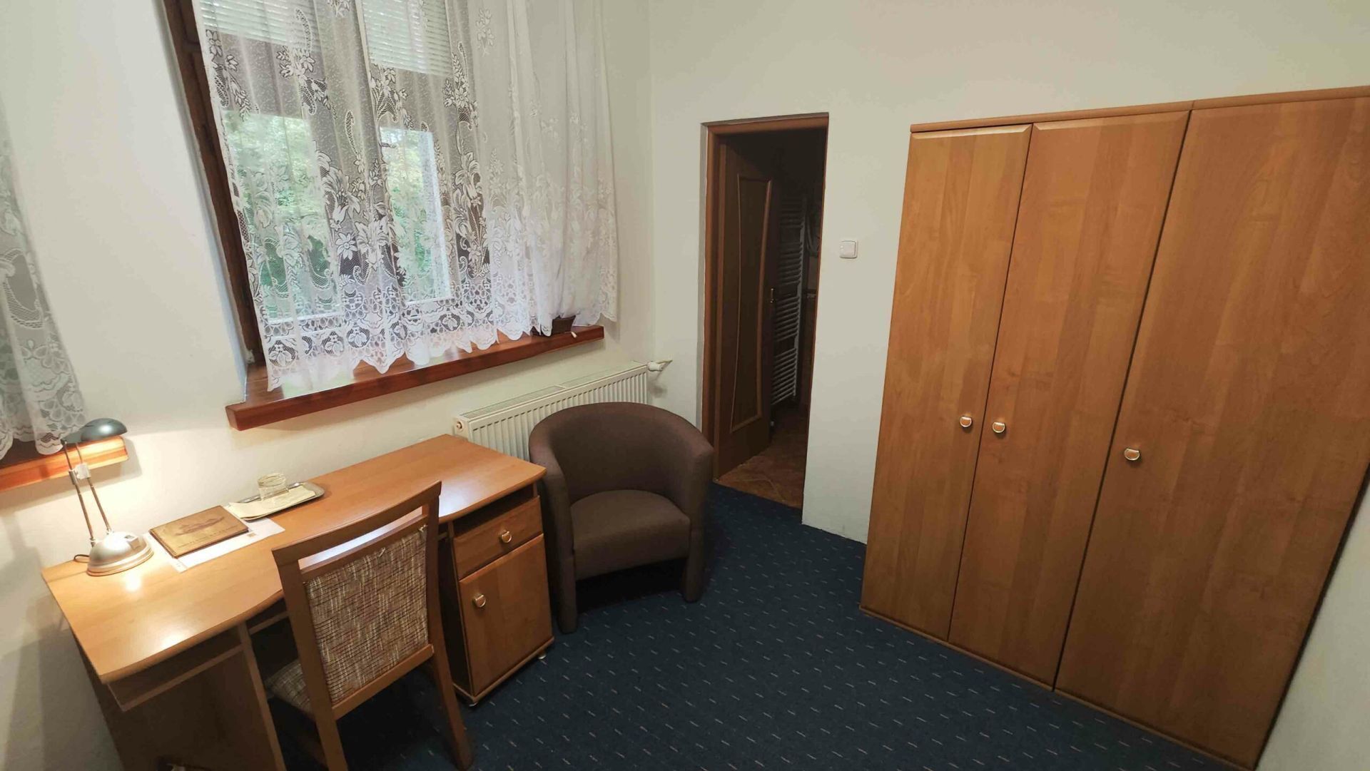 Une pièce avec un bureau en bois, une chaise, une armoire et une fenêtre ornée de rideaux en dentelle.