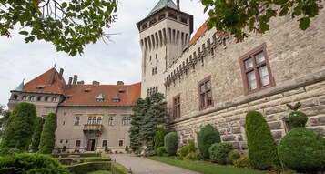 Um edifício histórico com uma torre, paredes de pedra e um jardim bem cuidado, com sebes e um caminho pavimentado.