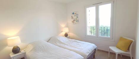 A bedroom with two beds, a nightstand with a lamp, a chair with a yellow cushion, and a window with a balcony view.