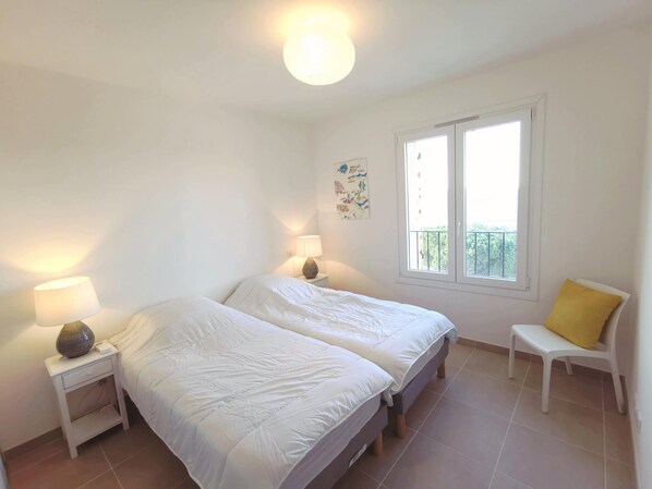 Une chambre avec deux lits, une table de chevet avec une lampe, une chaise avec un coussin jaune et une fenêtre donnant sur un balcon.