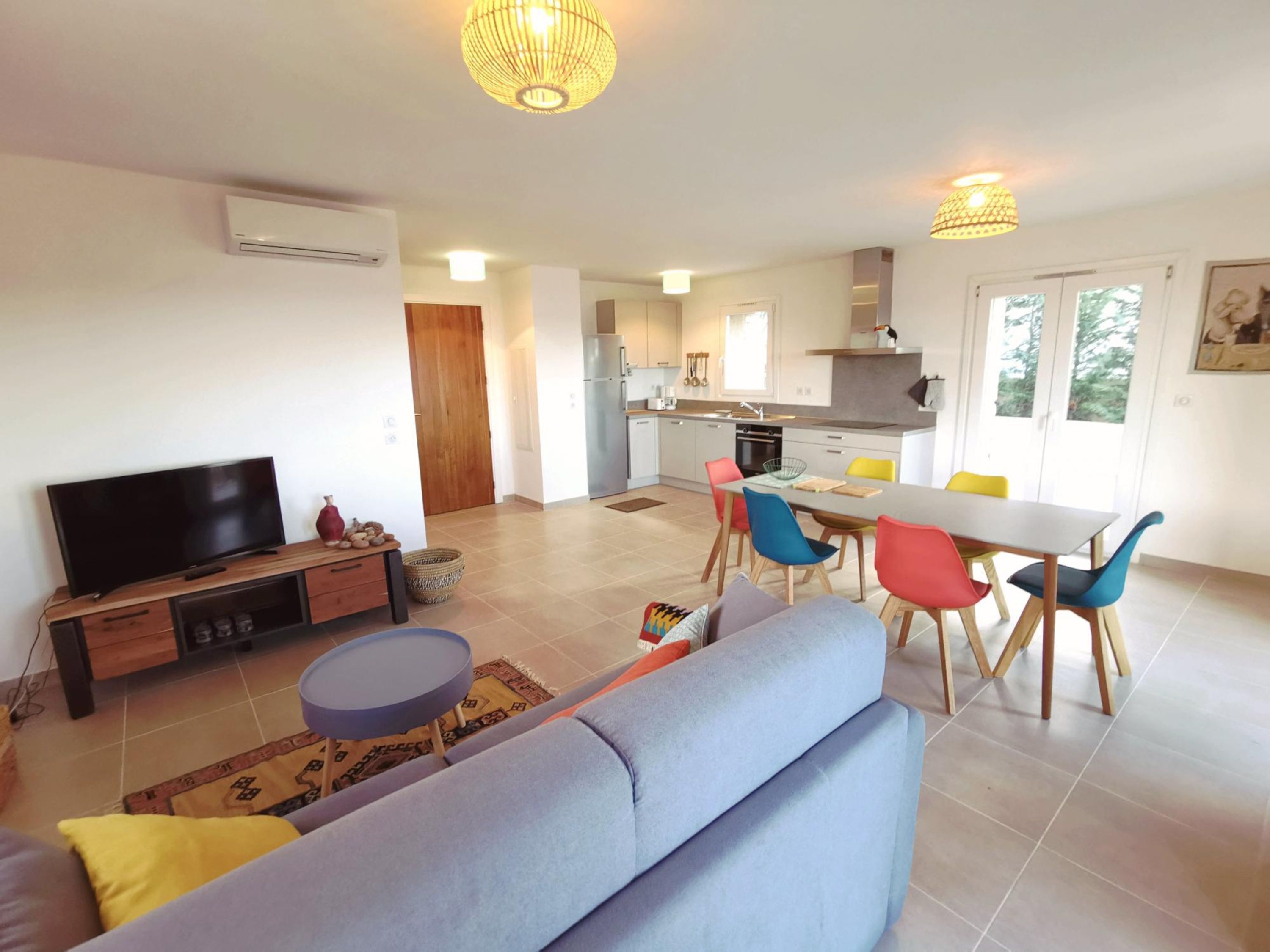 Una sala moderna con televisor de pantalla plana, un sofá, una mesa de comedor con sillas coloridas y una cocina al fondo.