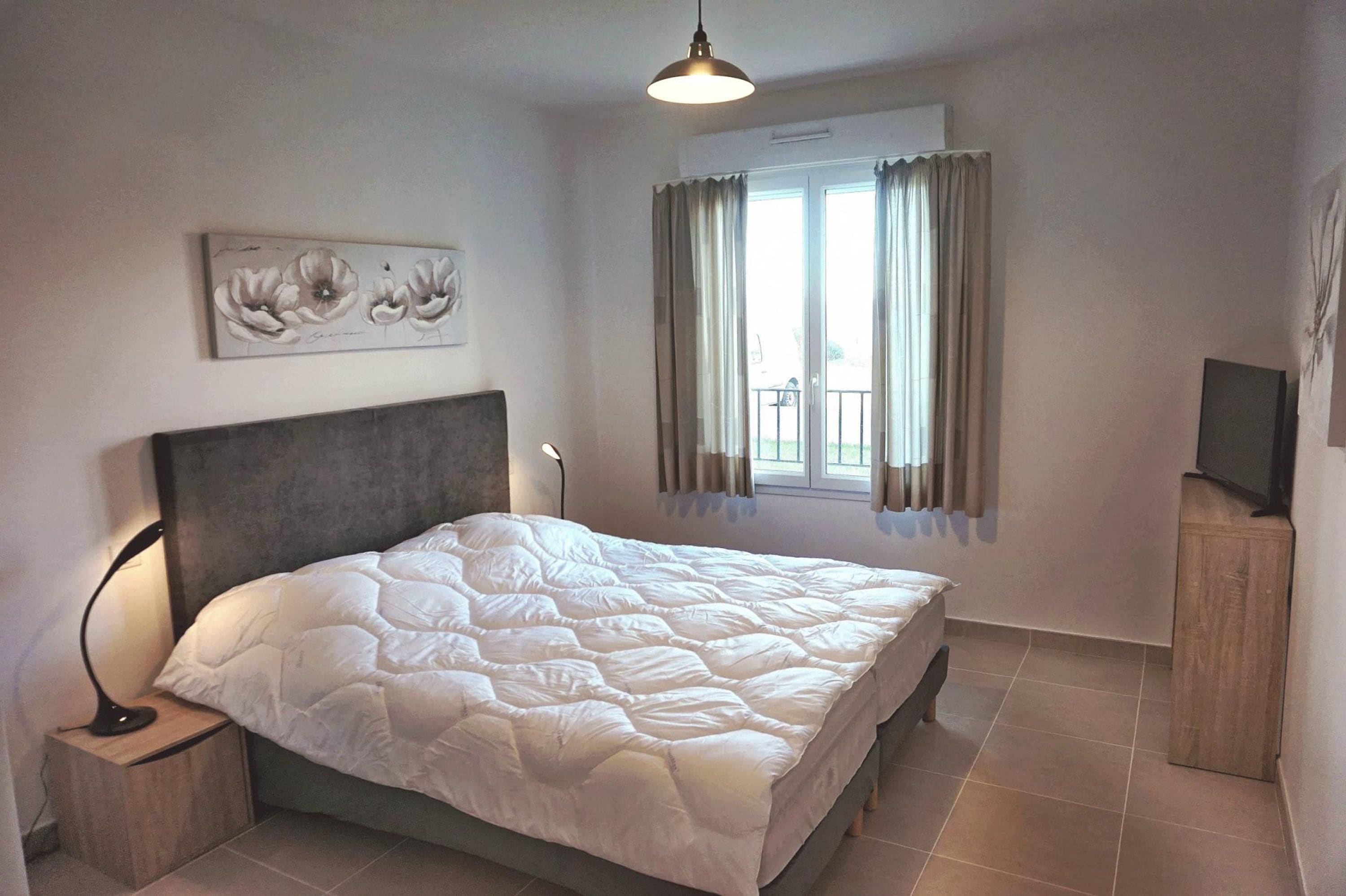 A bedroom with a bed, a nightstand, a TV, a window with curtains, and a wall-mounted artwork.