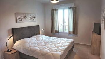 A bedroom with a bed, a nightstand, a TV, a window with curtains, and a wall-mounted artwork.
