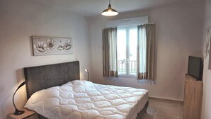 A bedroom with a bed, a nightstand, a TV, a window with curtains, and a wall-mounted artwork. - Mor102 - Appartement vue mer - 100m de la Plage (Saint-Florent)