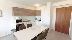 Een moderne keuken met witte kastjes, een magnetron, een oven en een spoelbak. Er staat een eettafel met stoelen en een deur aan de rechterkant.