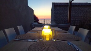 A wooden dining table with woven placemats, set for four, under a sunset. - Mor310-t2 Neuf vue mer Jardin Jacuzzi 100m Plage (Saint-Florent)