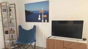 A room with a blue chair, a TV on a wooden cabinet, a bookshelf, and a painting of a landscape. - Mor310-t2 Neuf vue mer Jardin Jacuzzi 100m Plage (Saint-Florent)