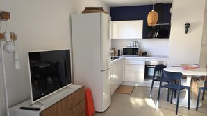 A compact kitchen with a refrigerator, microwave, and stove. There is a dining table with chairs and a TV on a cabinet. - Mor310-t2 Neuf vue mer Jardin Jacuzzi 100m Plage (Saint-Florent)