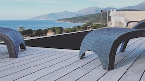 A deck with two wicker chairs, a white dog statue, and a view of the sea and mountains. - Mor405 - T3 Neuf vue mer Panoramique Avec Piscine (Saint-Florent)