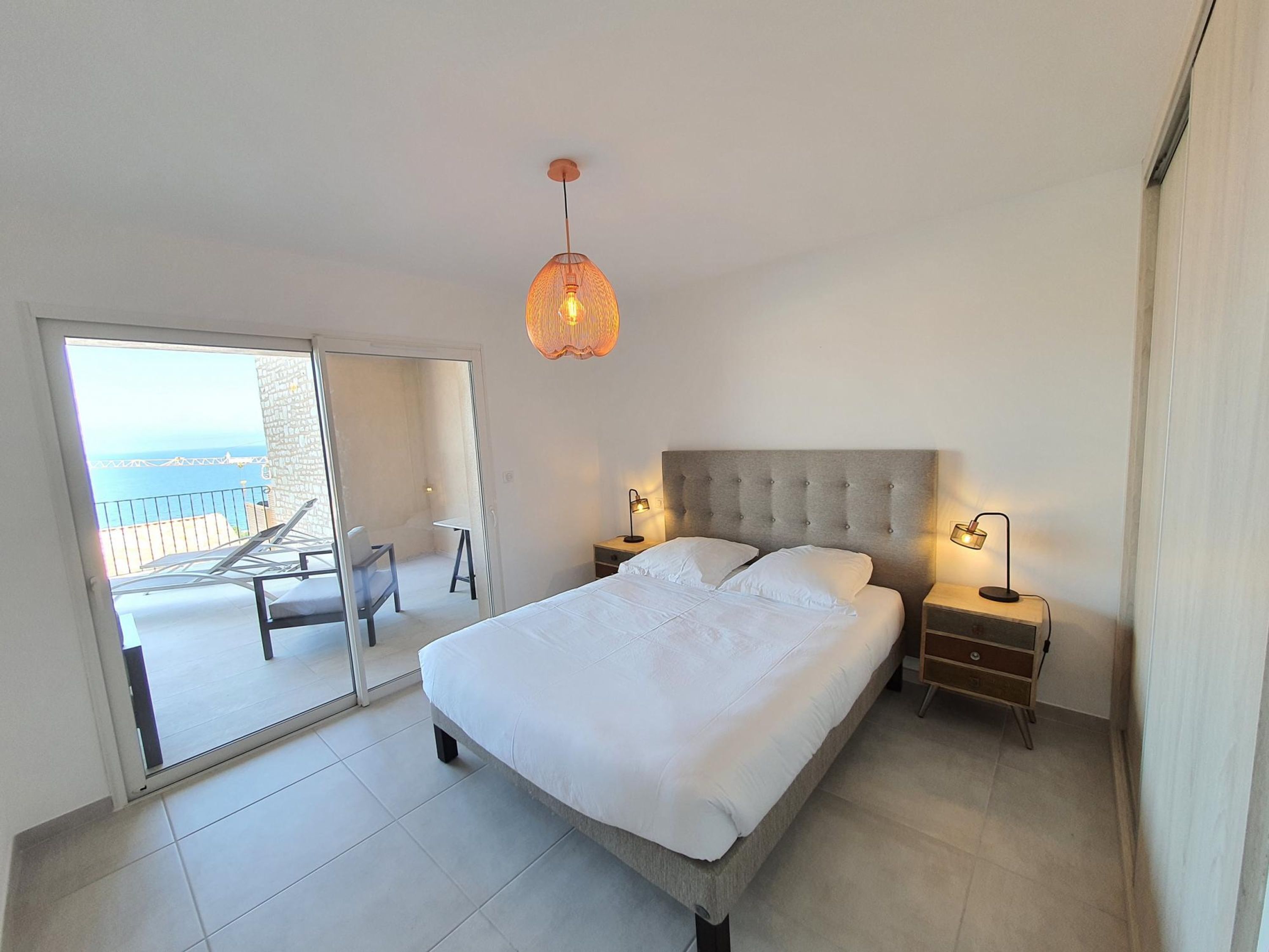 A bedroom with a bed, bedside tables, and a hanging light fixture. There is a sliding glass door leading to a balcony with a chair and a table.