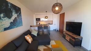 A modern living room with a black sofa, a television, and a dining area with a kitchen in the background. - Mor205 T2 Neuf vue mer acc s Plage (Saint-Florent)