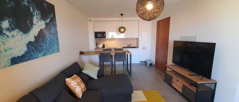 A modern living room with a black sofa, a television, and a dining area with a kitchen in the background.