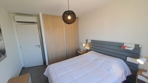 A bedroom with a bed, a wardrobe, a bedside table with a lamp, and a hanging light fixture. - Mor205 T2 Neuf vue mer acc s Plage (Saint-Florent)