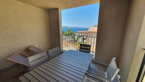 A balcony with a wooden table and chairs, a small table with a chair, and a view of a coastal area with buildings and mountains in the distance. - Mor205 T2 Neuf vue mer acc s Plage (Saint-Florent)