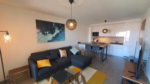 A modern living room with a sofa, coffee table, and a painting on the wall. There is a kitchen area with a dining table and chairs. - Mor205 T2 Neuf vue mer acc s Plage (Saint-Florent)
