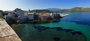A coastal town with historic buildings, clear blue waters, and mountains in the background.