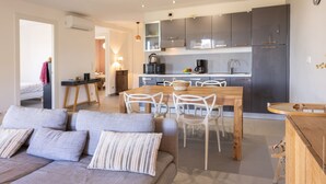 Una cocina moderna con gabinetes oscuros, una mesa de comedor de madera y sillas blancas. En la sala de estar, hay un sofá con almohadones a rayas.