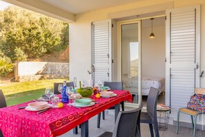 An outdoor dining area with a table set for four, chairs, a small table with a chair, and a bed visible through an open door.