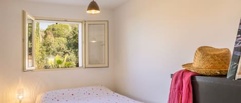 Une chambre à coucher comprenant un lit, une table de chevet, une commode, une fenêtre donnant sur un espace verdoyant et un luminaire suspendu.