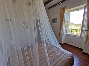 Un dormitorio con una cama cubierta por un dosel blanco translúcido, una ventana con vistas a un paisaje y un suelo de baldosas.