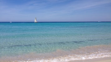 A beach with clear turquoise water and a sailboat in the distance.