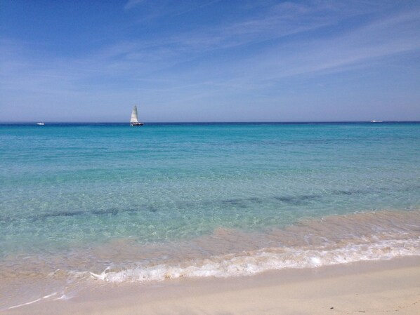 Una playa con agua turquesa cristalina y un velero a lo lejos.