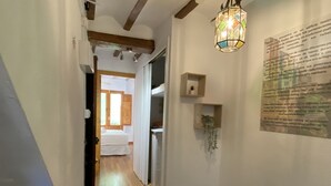 Ein Korridor mit Holzböden, weißen Wänden und freiliegenden Holzbalken. Eine Pendelleuchte und ein an der Wand befestigtes Regal mit einer Pflanze sind vorhanden.