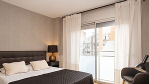 A modern hotel room with a large bed, a nightstand with a lamp, a chair, and a view of the city through a sliding glass door.