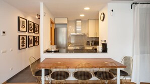 A modern kitchen with a stainless steel refrigerator, a built-in oven, and a microwave. There is a wooden dining table with four wireframe chairs.