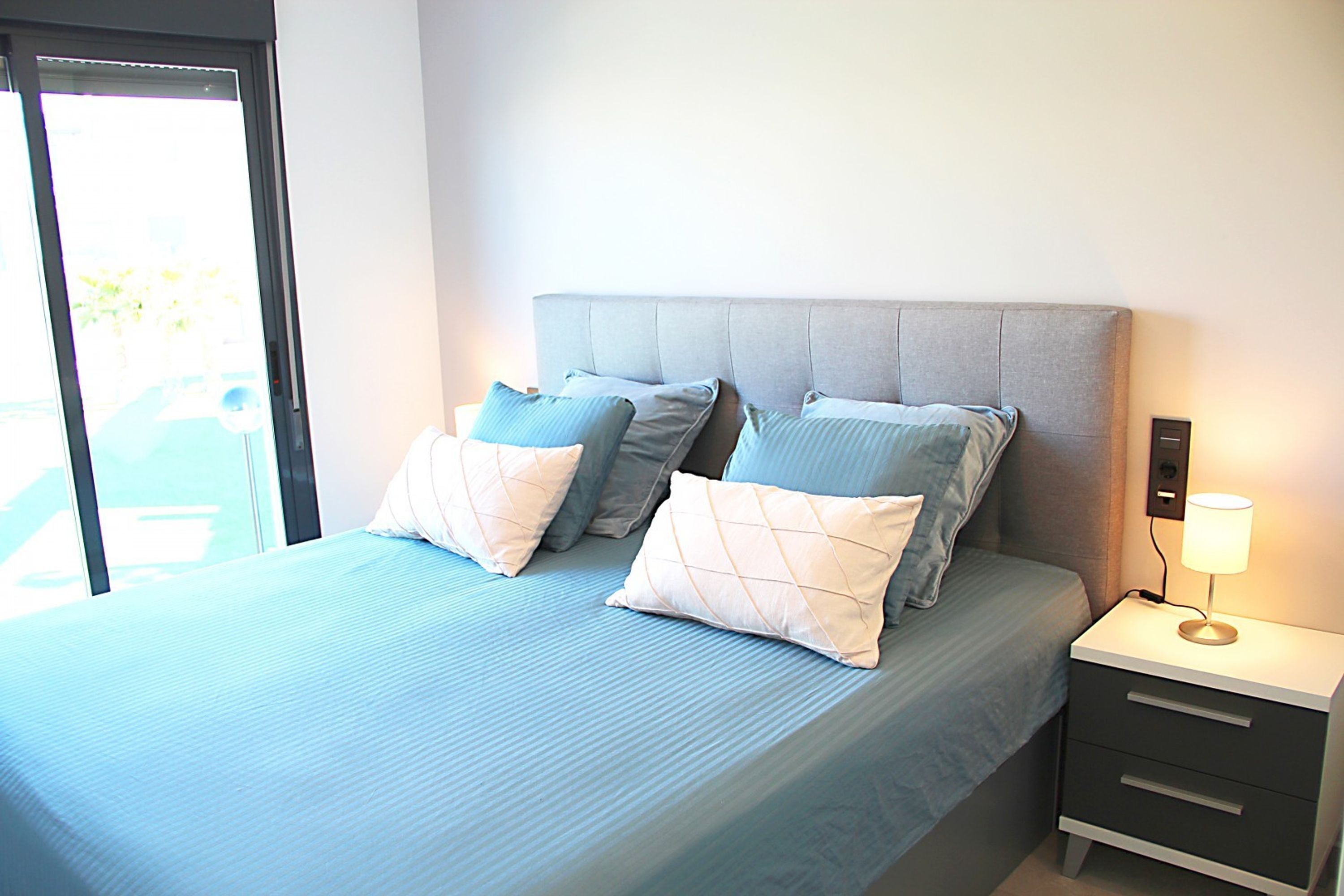 A neatly made bed with a grey headboard, two bedside tables with lamps, and a view of the outdoors through a sliding door.