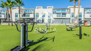 A modern outdoor gym area with exercise equipment, palm trees, and a building with balconies in the background. - Casa Rosa in Guardamar (Guardamar del Segura)