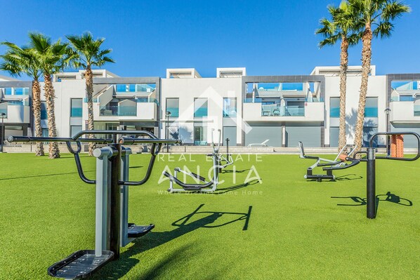 A modern outdoor gym area with exercise equipment, palm trees, and a building with balconies in the background.